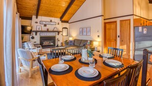 Un comedor con una mesa de madera dispuesta para cuatro, una chimenea y una sala de estar con un sofá y un televisor.