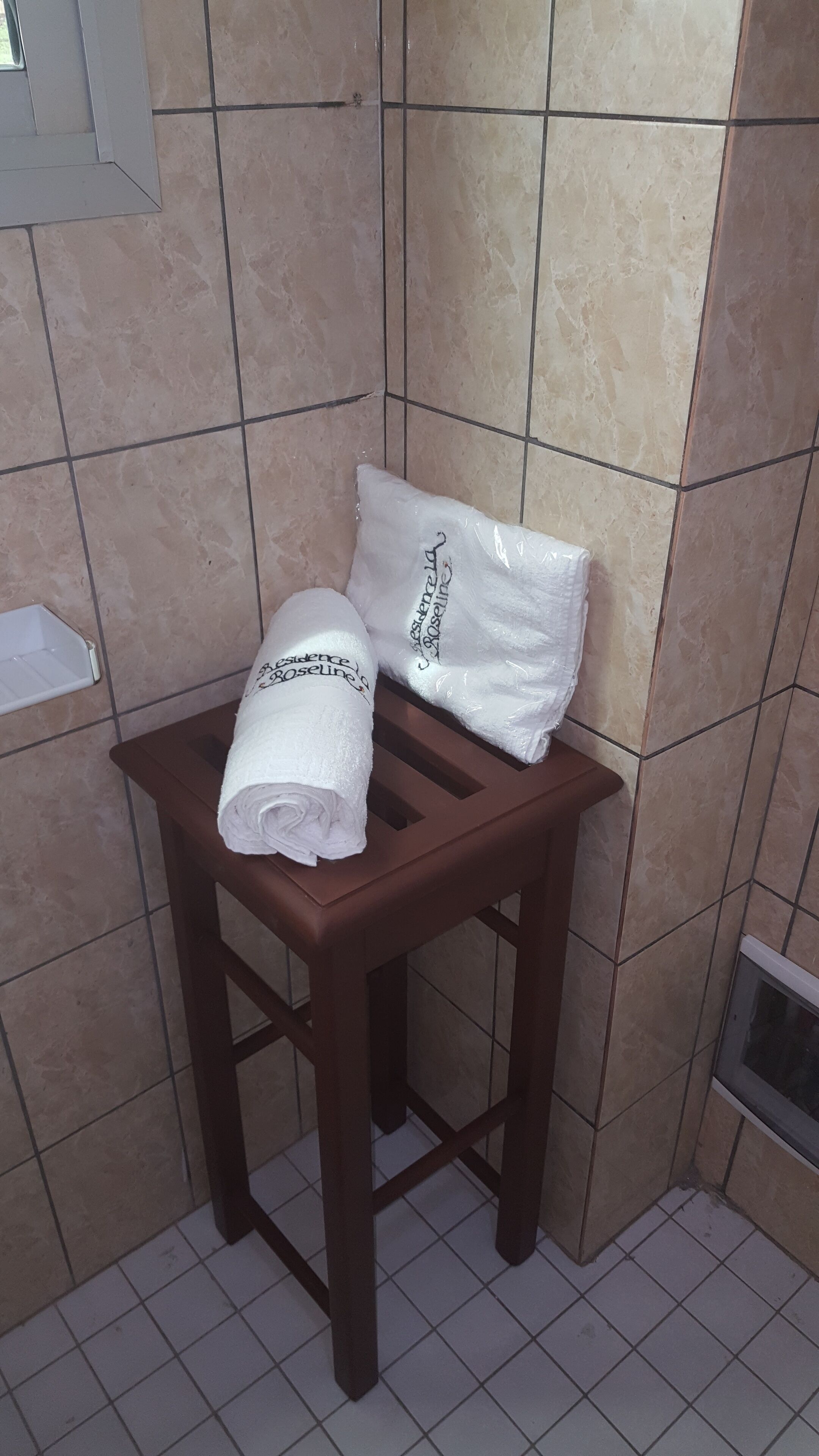 Een badkamer met beige tegels, een houten kruk en twee opgerolde handdoeken met de merknaam 'Rosaline'.