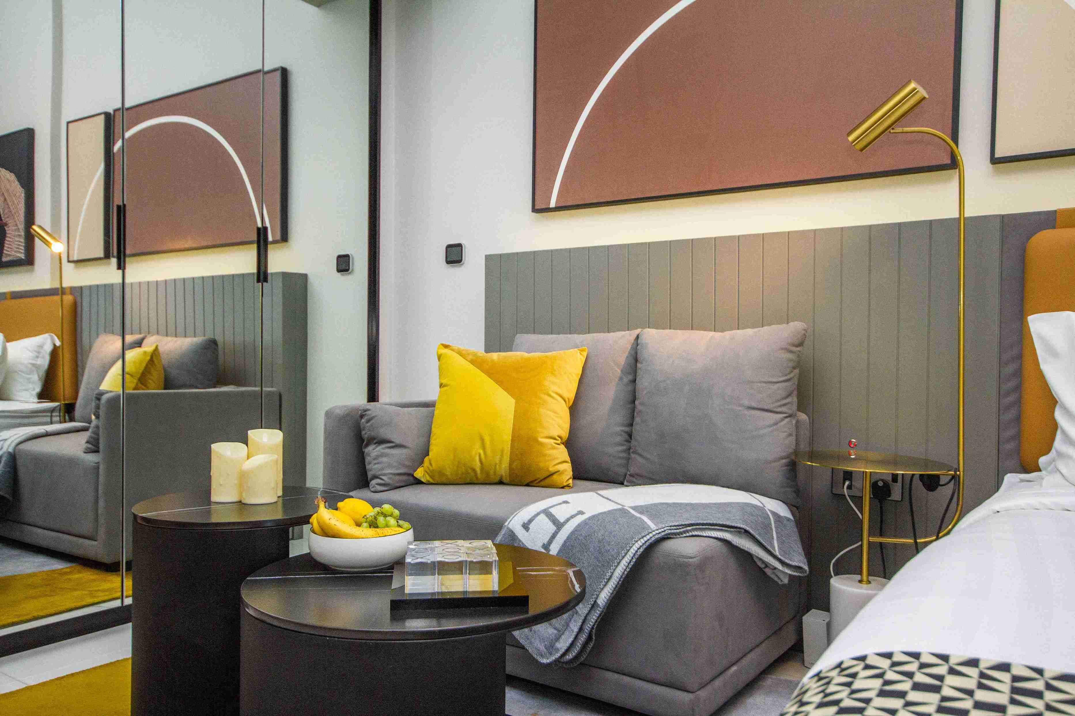 A modern hotel room with a grey sofa, a small round table with a bowl of fruit, a bedside table with a lamp, and a bed with a patterned bedspread.