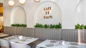 A restaurant interior with a modern design, featuring a seating area with tables set for dining, a wall with the text "CAFE DE PARIS", and green plants along the lower part of the wall. - Studio Canal & City View, Pool & Gym Access, MAG 318 Business Bay (Business Bay)