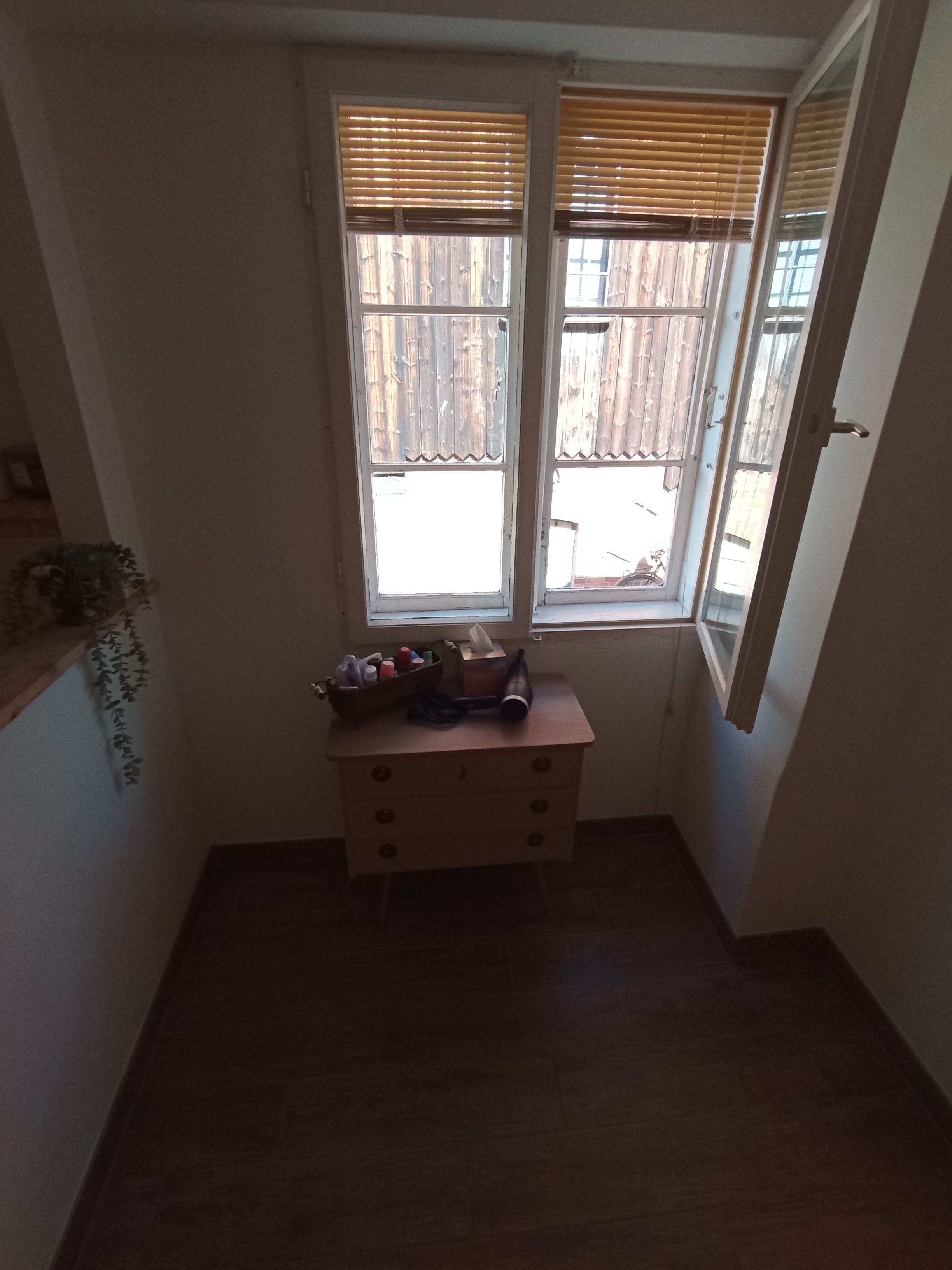 Ein schmaler Raum mit Holzboden, einem kleinen Holzschrank mit diversen Gegenständen und zwei Fenstern mit Holzjalousien.