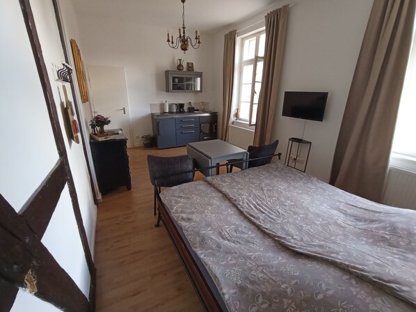 A bedroom with a bed, a desk, a chair, a television, and a kitchenette.