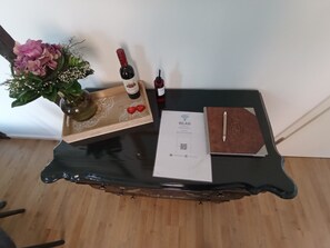 Una pequeña mesa con una bandeja que sostiene una botella de vino, un florero con flores, una copa de vino, un folleto y un código QR.