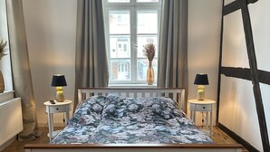 Een slaapkamer met een houten bed, een raam met gordijnen, twee nachtkastjes met lampen en uitzicht op gebouwen.