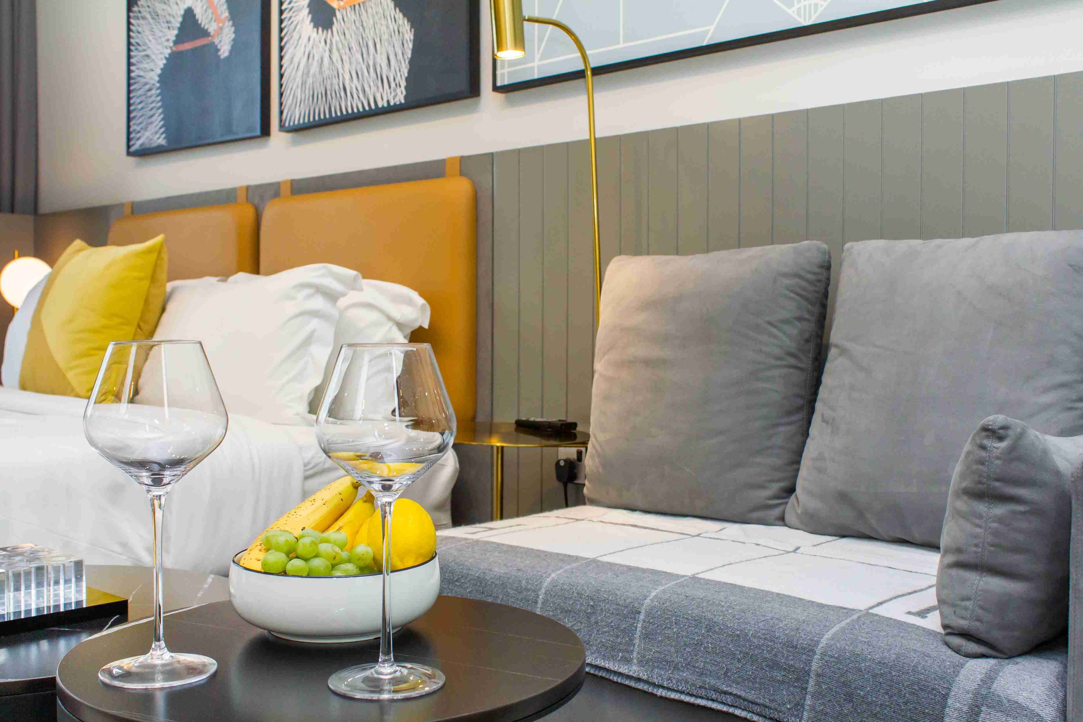 Et moderne hotelværelse med en seng, en grå sofa og et bord med to vinglas samt en skål med frugt.