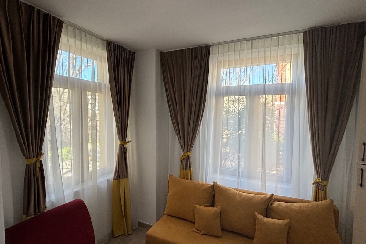 A room with a sofa, red chair, and curtains tied back to reveal windows.