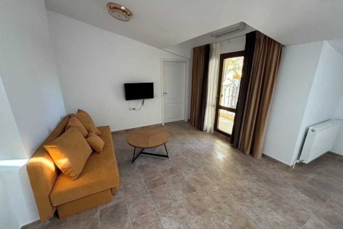 Un salon comprenant un canapé, une petite table, un téléviseur à écran plat fixé au mur et une fenêtre avec des rideaux.