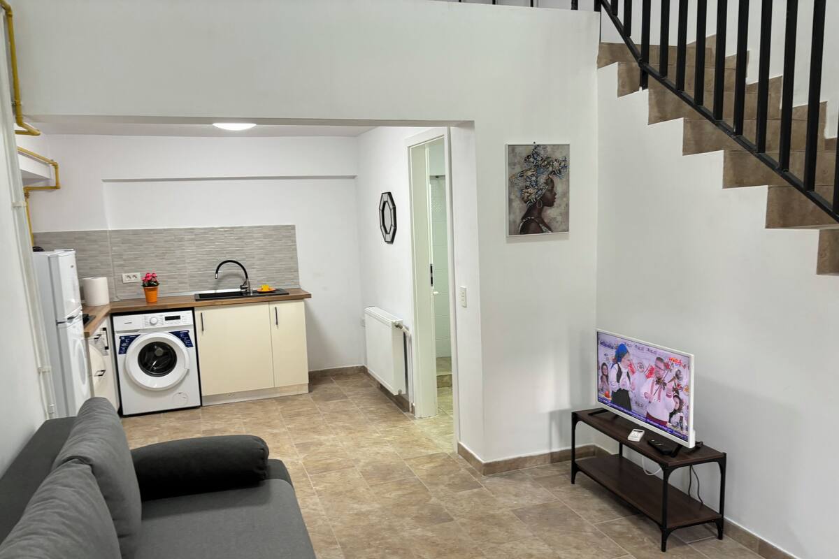 A modern living room with a grey sofa, a kitchen area with a washing machine, a dining table with a TV, and a staircase.