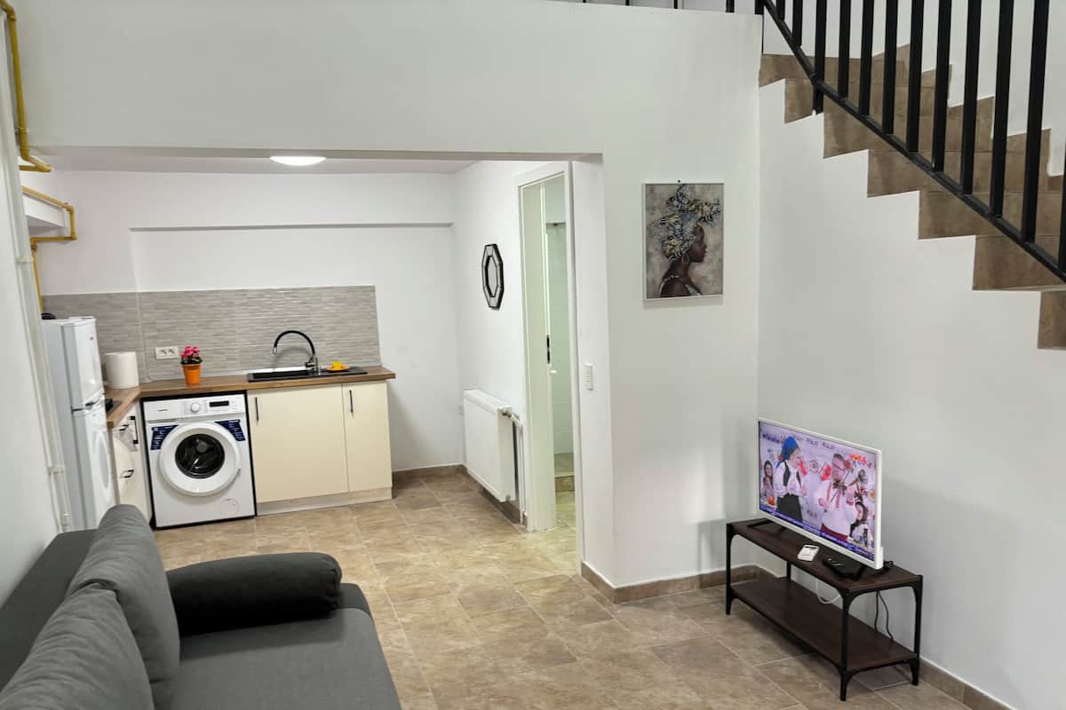 A modern living room with a grey sofa, a kitchen area with a washing machine, a dining table with a TV, and a staircase.