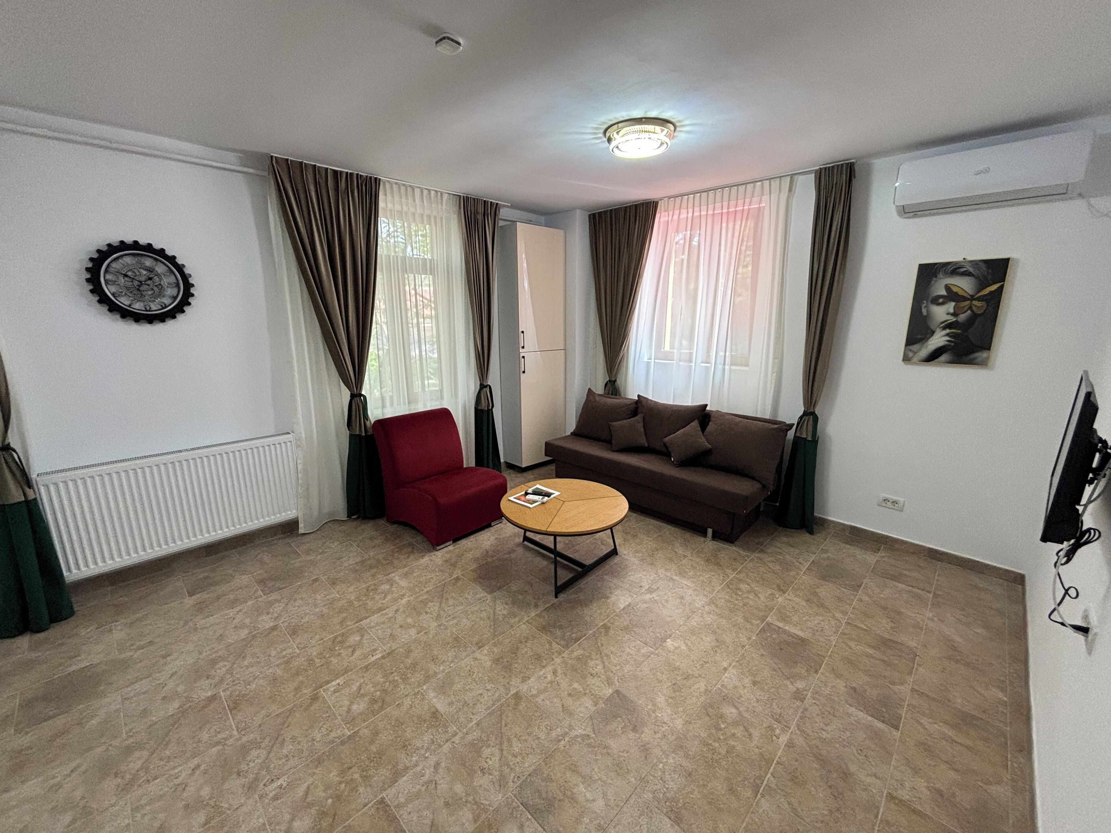 Una sala de estar con una silla roja, un sofá marrón, una mesa de centro redonda y un reloj en la pared.