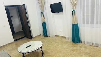 A room with a tiled floor, a round table with a remote control, a TV mounted on the wall, and a door leading to another room.