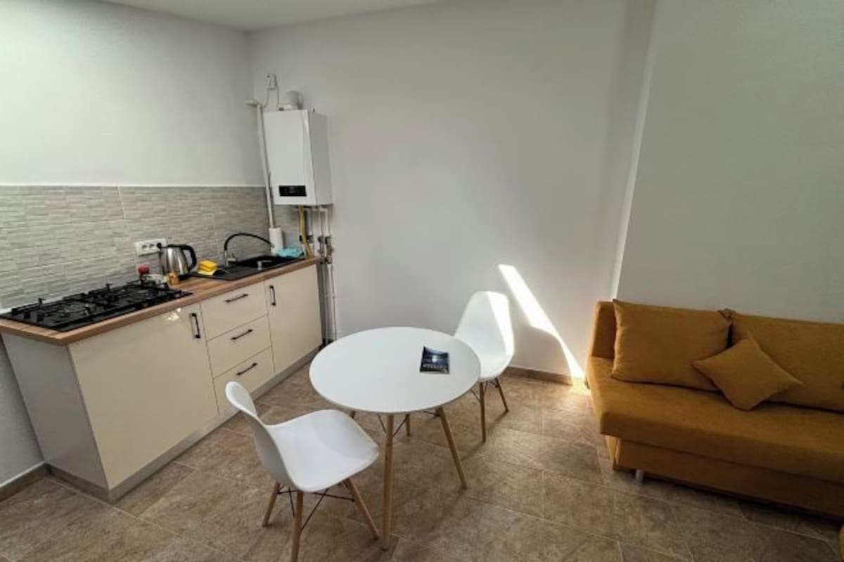 A compact kitchen with a gas stove, white cabinets, and a white refrigerator. There is a round table with two white chairs and a mustard-colored sofa against the wall.