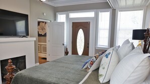 Une chambre à coucher avec un grand lit, un téléviseur placé au-dessus d’une cheminée et une porte ornée d’un panneau de verre décoratif.