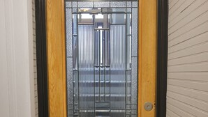 Une porte en bois, dotée d’un panneau de verre et d’une grille métallique, est installée dans un couloir aux murs blancs et au plancher en bois.