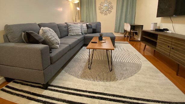 Un salon moderne avec un canapé d’angle gris, une table basse en bois, un tapis à rayures, un téléviseur à écran plat et une petite table d’appoint.