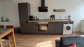 Una cocina moderna con horno integrado, microondas y lavadora. Cuenta con una mesa de comedor con sillas y un sofá con cojín.