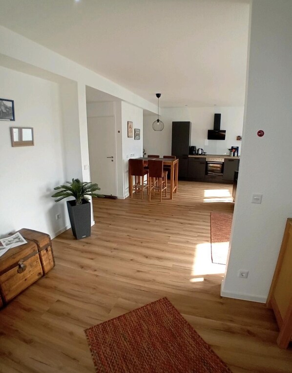 Ein geräumiger Wohnbereich mit Holzboden, einem Esstisch mit Stühlen, einer Küche im Hintergrund und einer Zimmerpflanze.