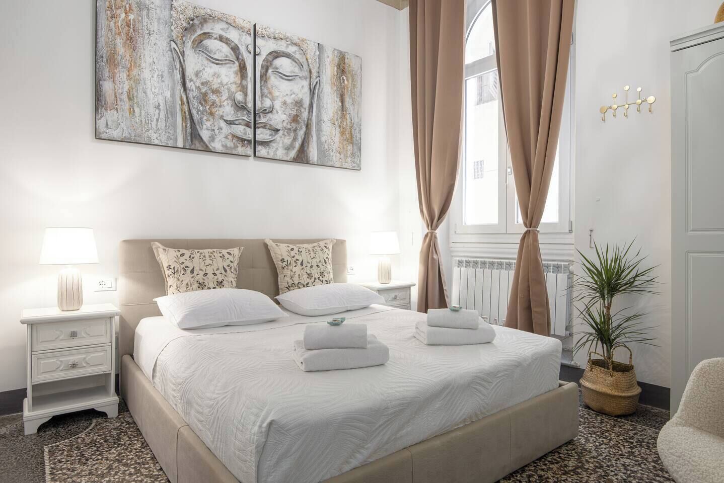 A neatly arranged bedroom with a large bed, two bedside tables with lamps, a wall-mounted artwork, and a window with curtains.