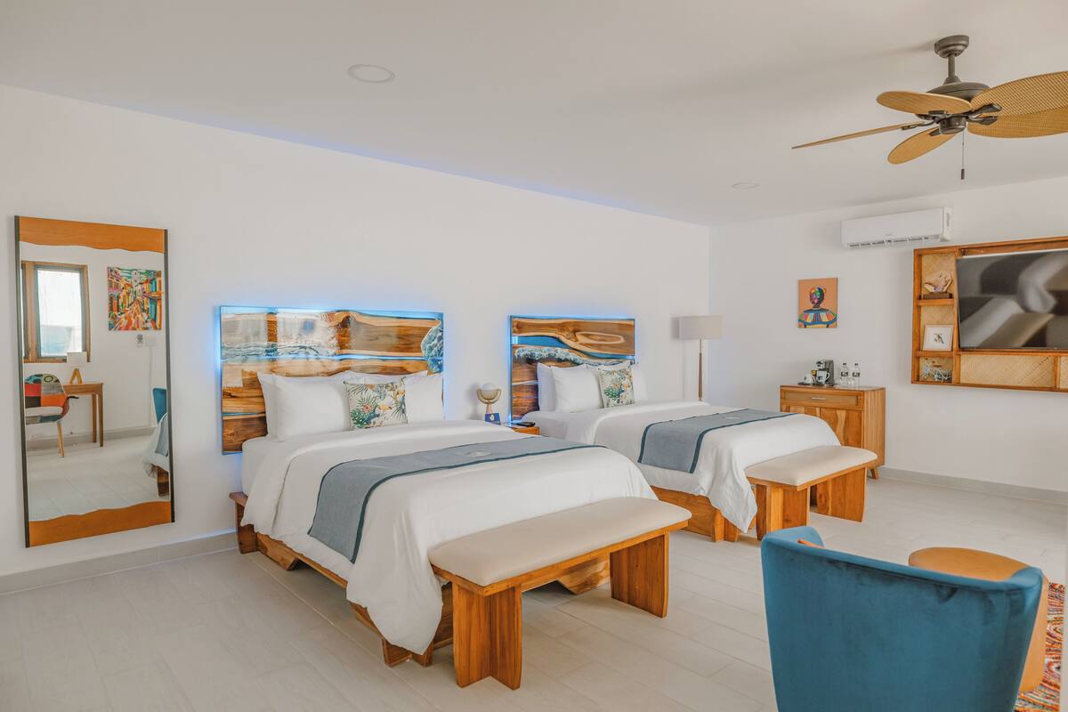 Habitación de hotel con dos camas, ventilador de techo, un espejo grande y obras de arte en las paredes.