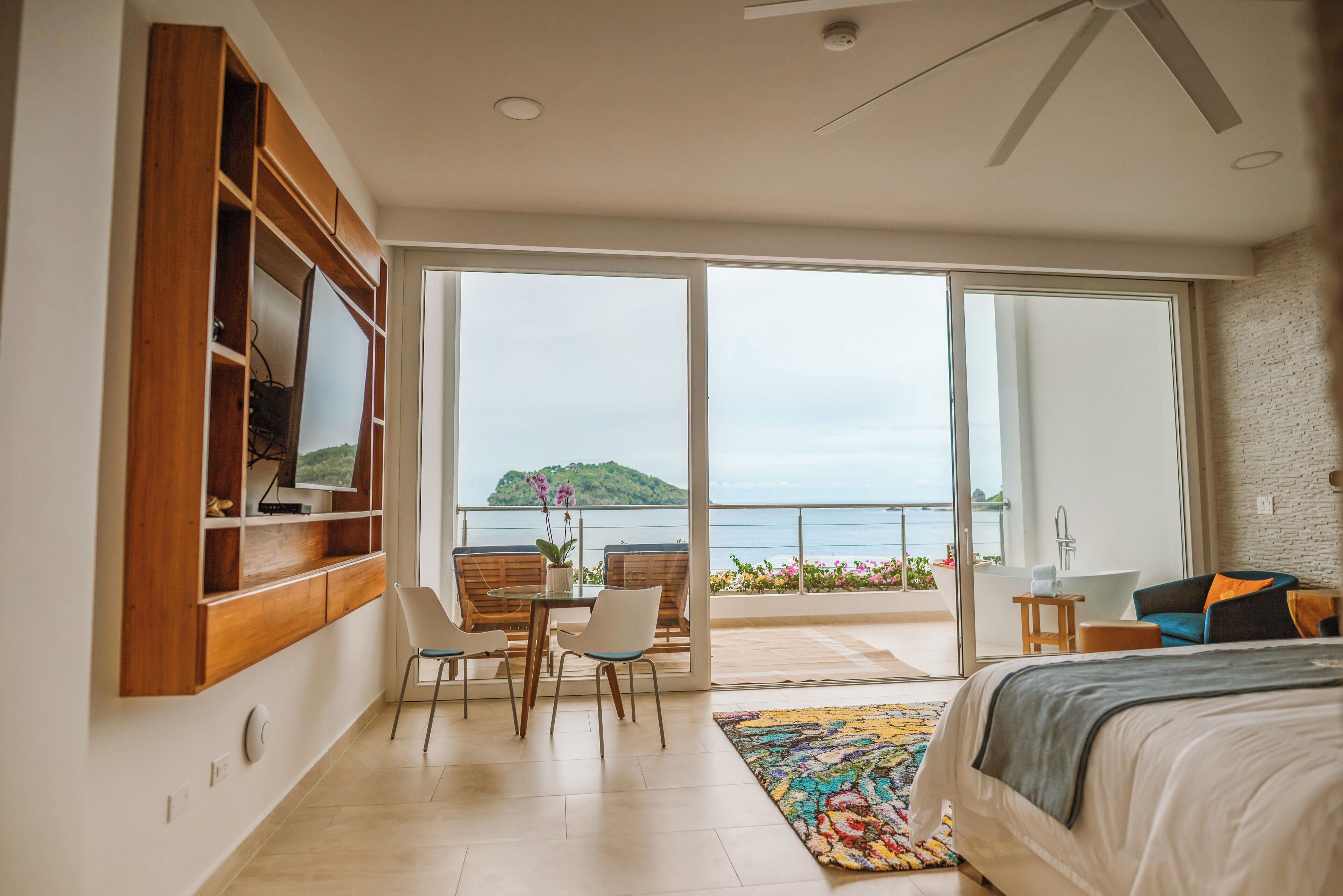 Un dormitorio con una cama grande, un escritorio, sillas, vista al mar y un balcón con bañera.