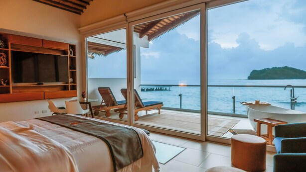 A bedroom with a bed, a TV, and a balcony with a view of the sea.