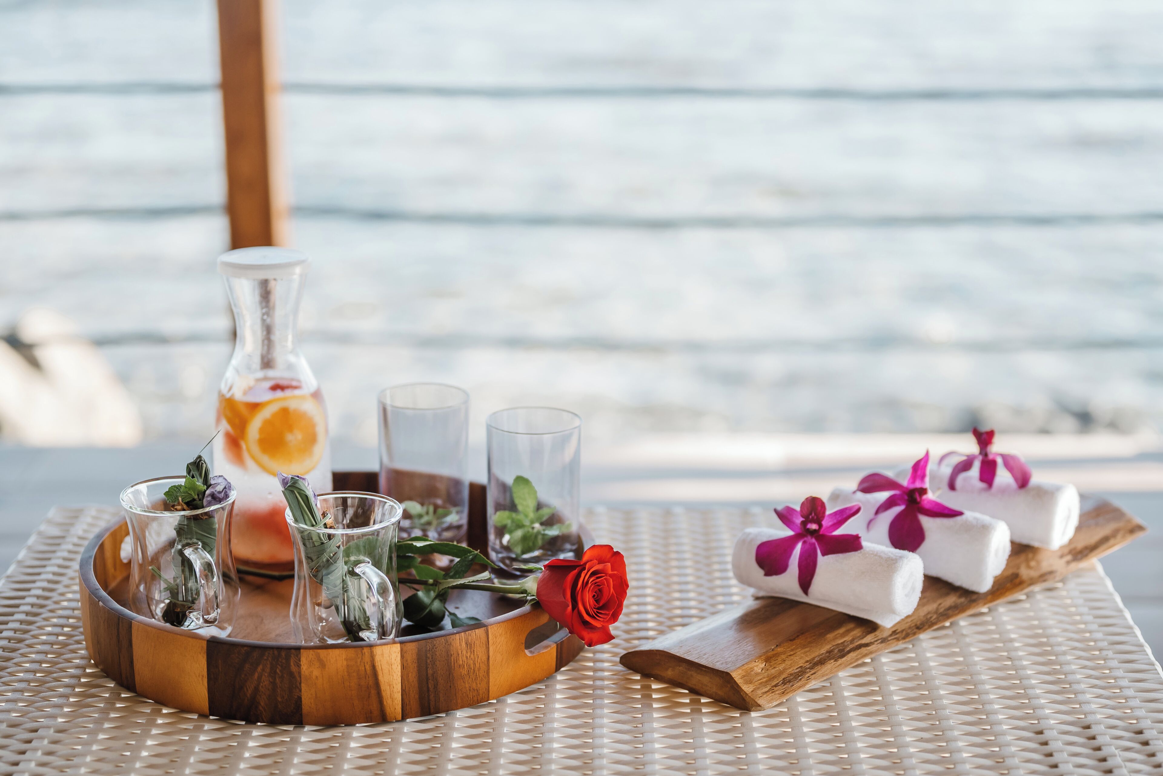 A wooden tray with a glass bottle, shot glasses, a red rose, and towels with purple flowers on a woven table.