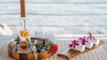 A wooden tray with a glass bottle, shot glasses, a red rose, and towels with purple flowers on a woven table.
