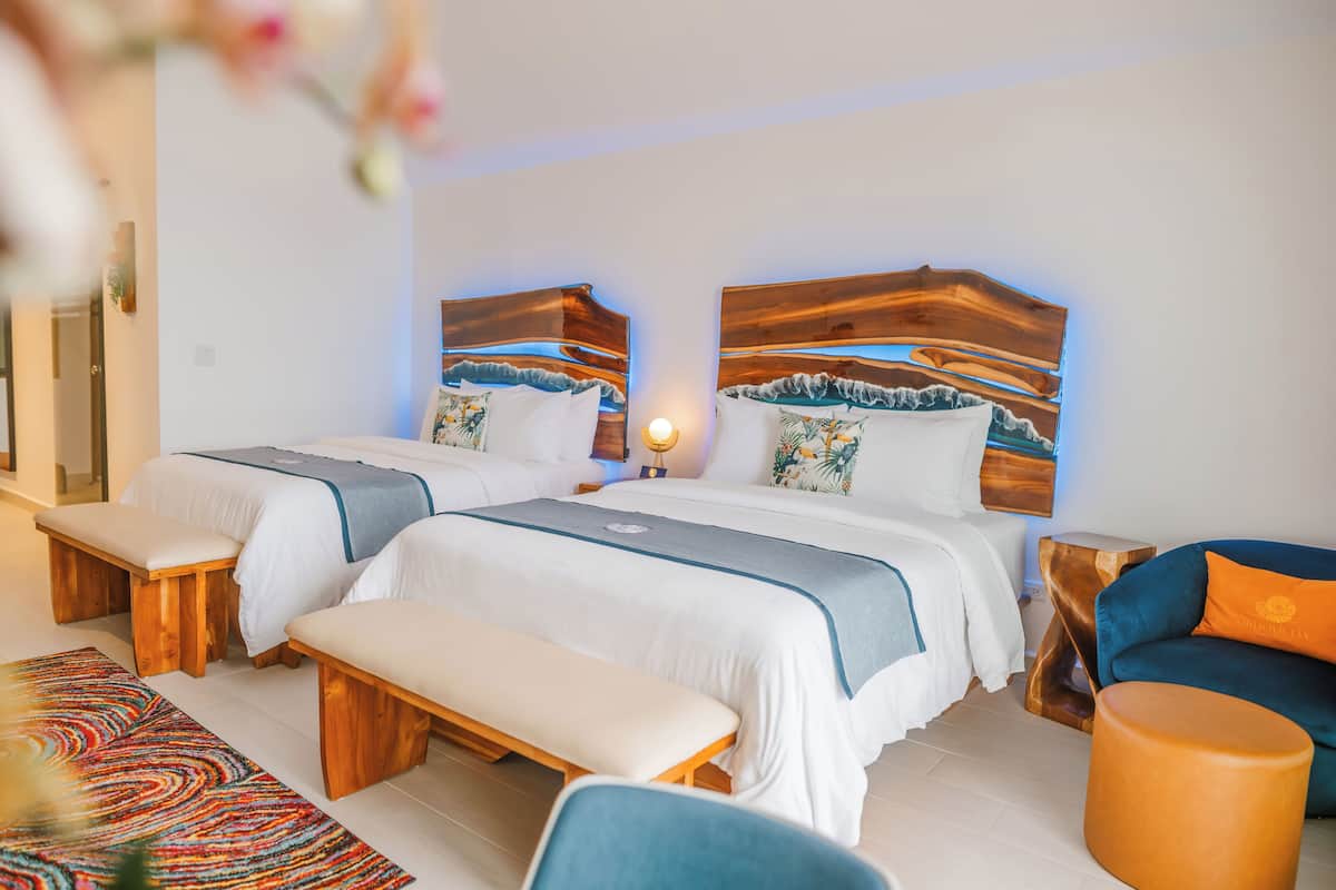 Habitación de hotel con dos camas, bancos de madera, una alfombra colorida y un sillón azul.