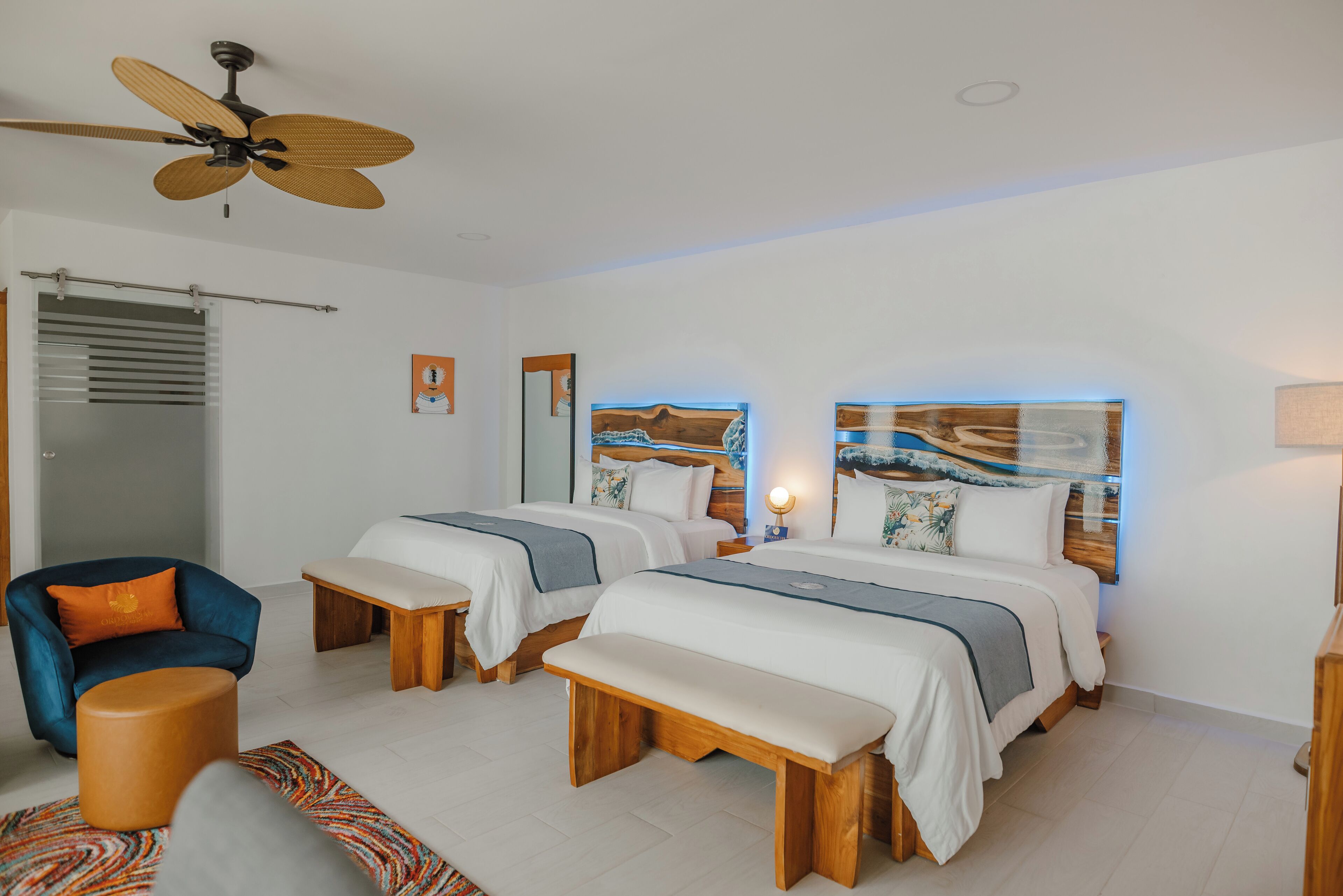 A hotel room with two beds, a ceiling fan, a chair, a bench, and artwork on the walls.