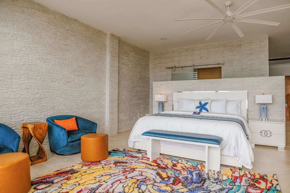 Un dormitorio con cama blanca, ventilador de techo, alfombra colorida y pared de ladrillo.