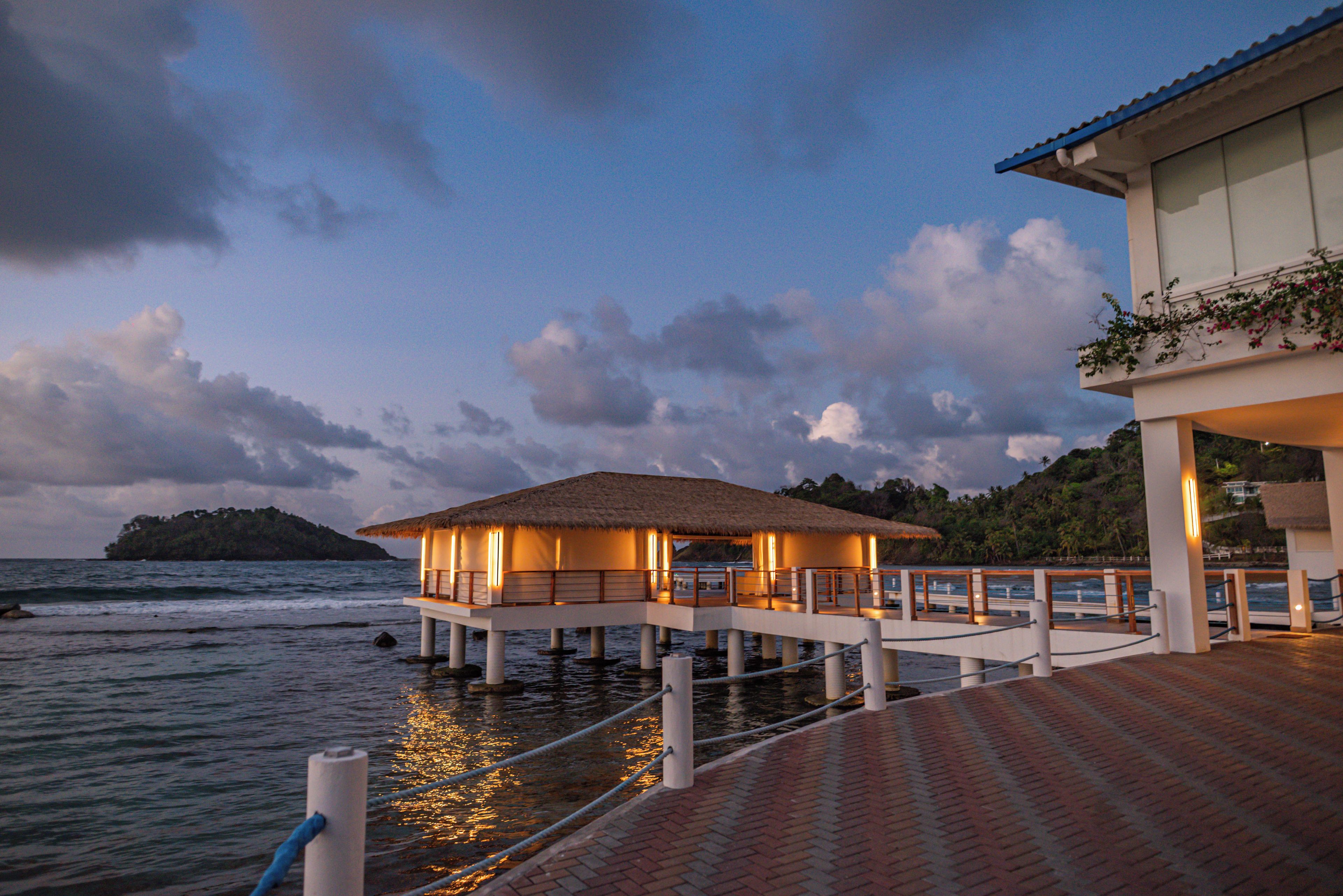 Bungalows auf Stelzen mit Reetdächern, verbunden durch Wege über dem Wasser, mit Blick auf das Meer und eine entfernte Insel.