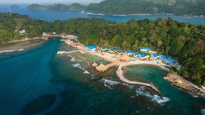 An aerial view of a coastal resort with multiple buildings, a beach, and surrounding forested hills.