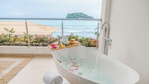 A bathtub with water, towels, and flowers on a balcony overlooking the sea.