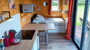 A compact cabin with a wooden interior, a bed with patterned bedding, a small kitchenette, and a window with a view of greenery.