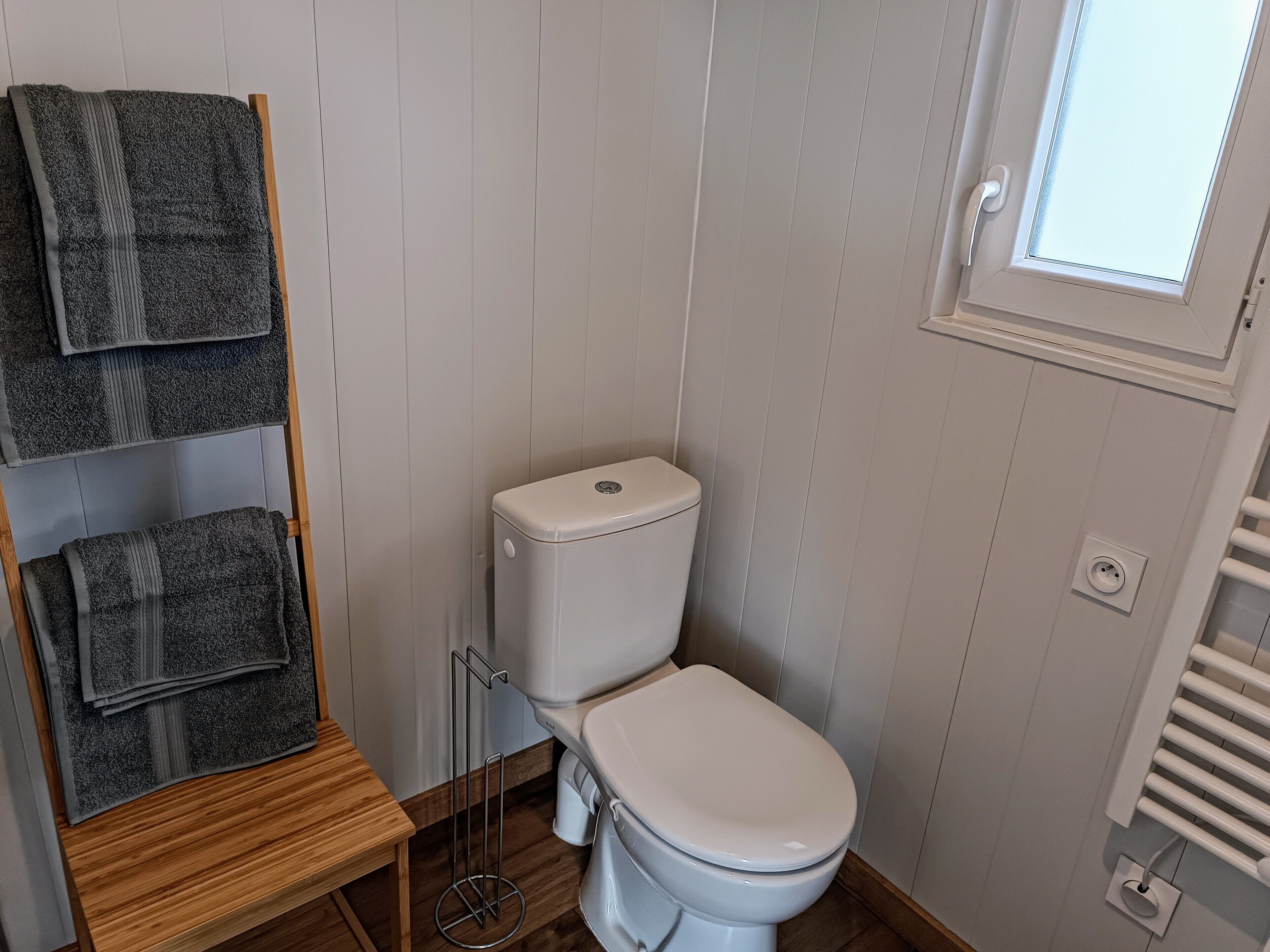 Et badeværelse med toilet, et håndklædestativ med foldede håndklæder, en træskammel og et vindue med udsigt.