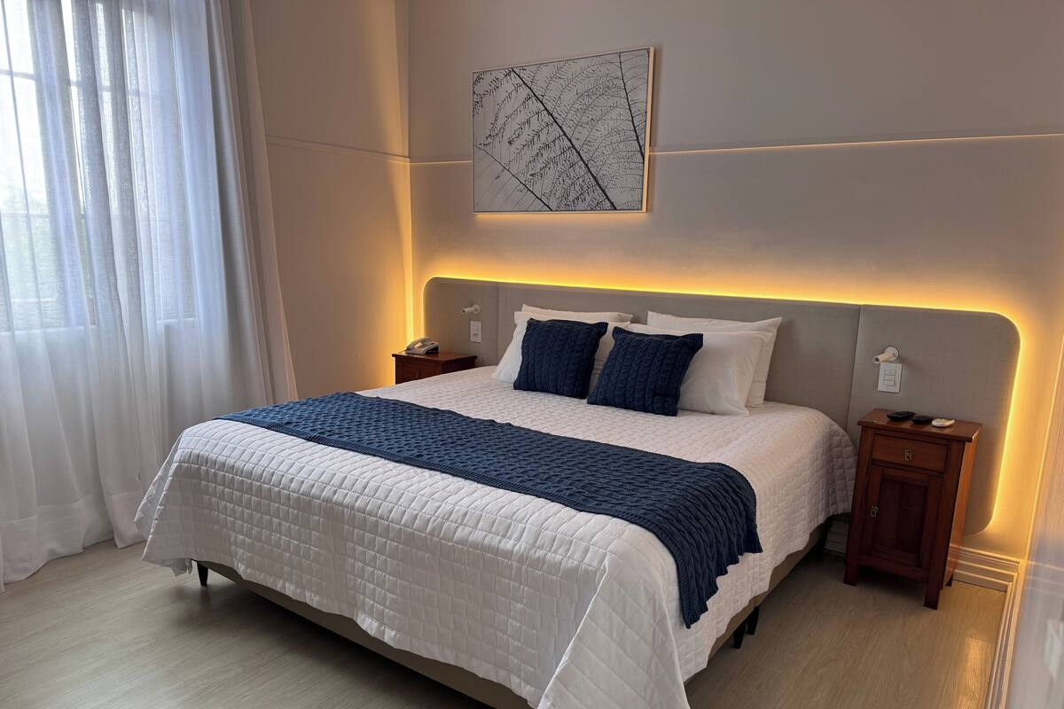 Una cama bien hecha con colcha blanca y azul, una mesita de noche con lámpara y una obra de arte en la pared.