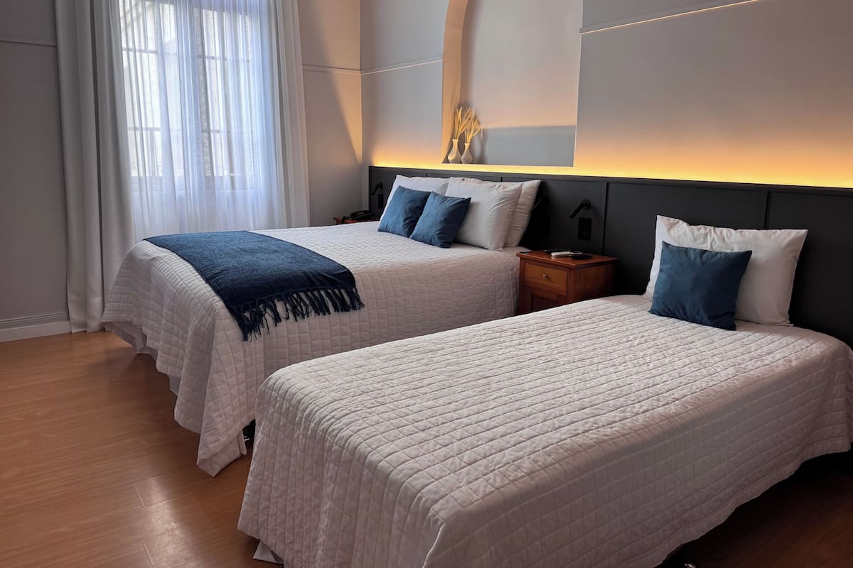 Habitación de hotel con dos camas, una mesita de noche, un espejo y una ventana con cortinas traslúcidas.