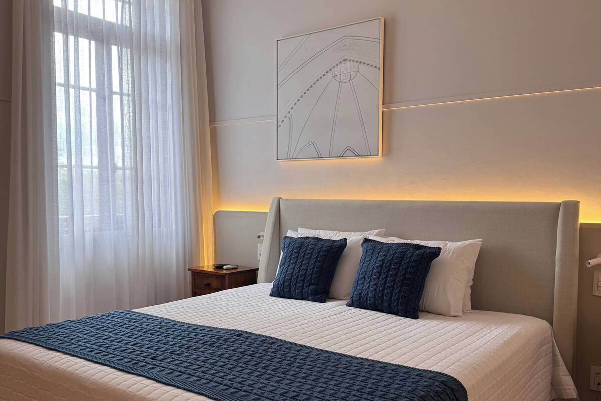 Una cama bien hecha con colcha blanca y azul, cabecera con lámpara, y un ventanal con cortinas traslúcidas.