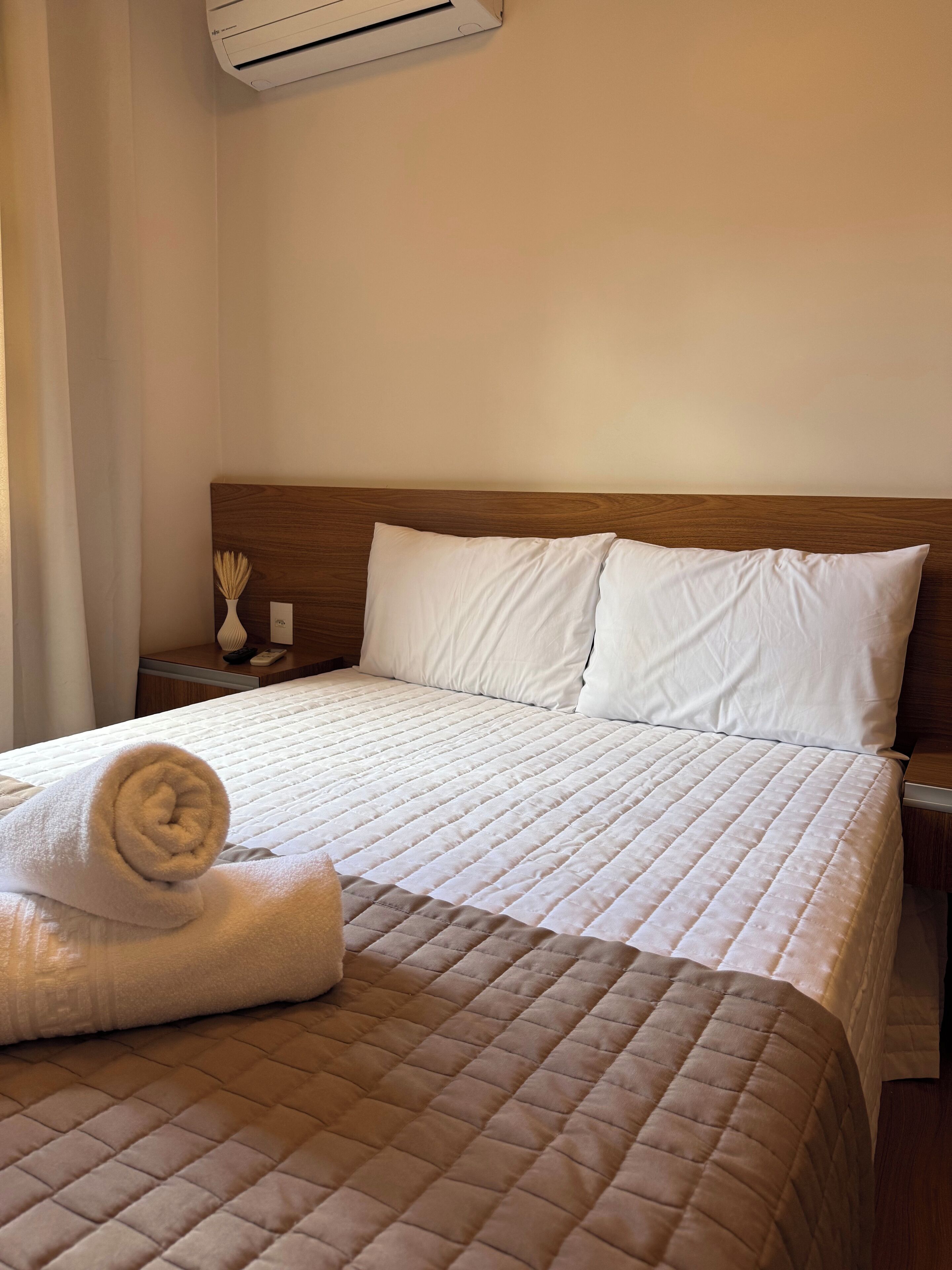 Ein ordentlich bezogenes Bett mit einer gesteppten braunen Decke, weißen Kissen und einem aufgerollten weißen Handtuch daneben.