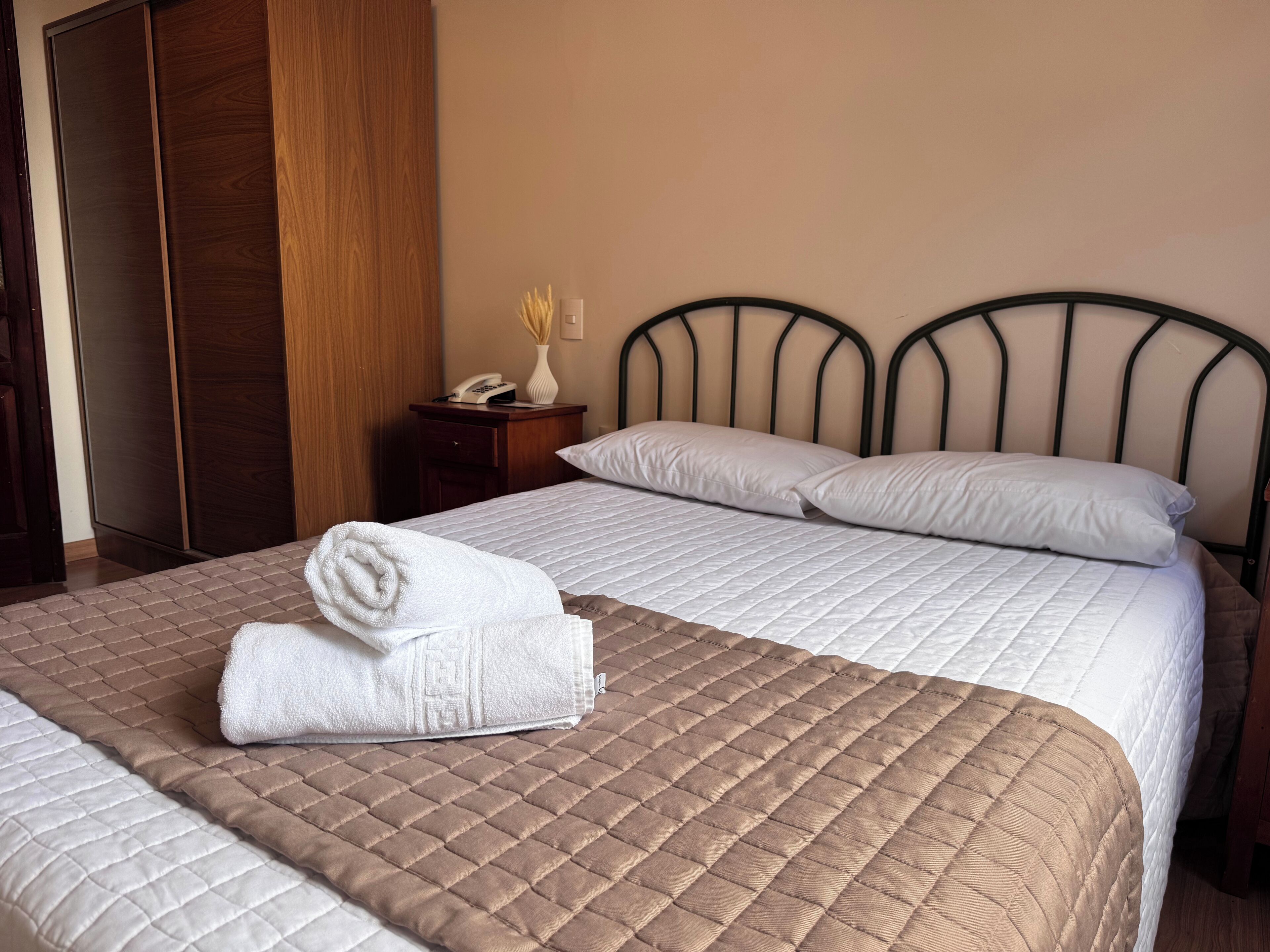 A hotel room with a bed, a wooden wardrobe, a nightstand with a phone, and a vase with dried flowers.