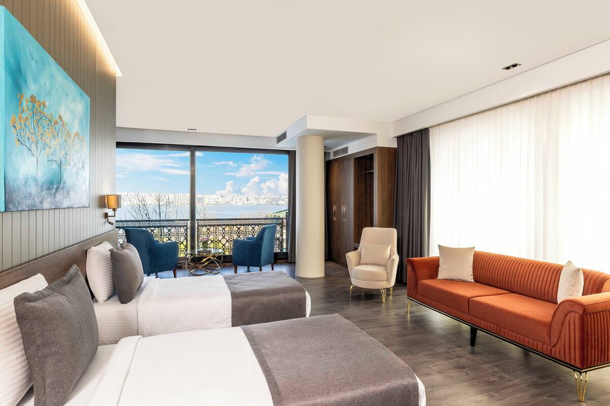 A modern hotel room with two beds, a sofa, and a balcony with a city view.