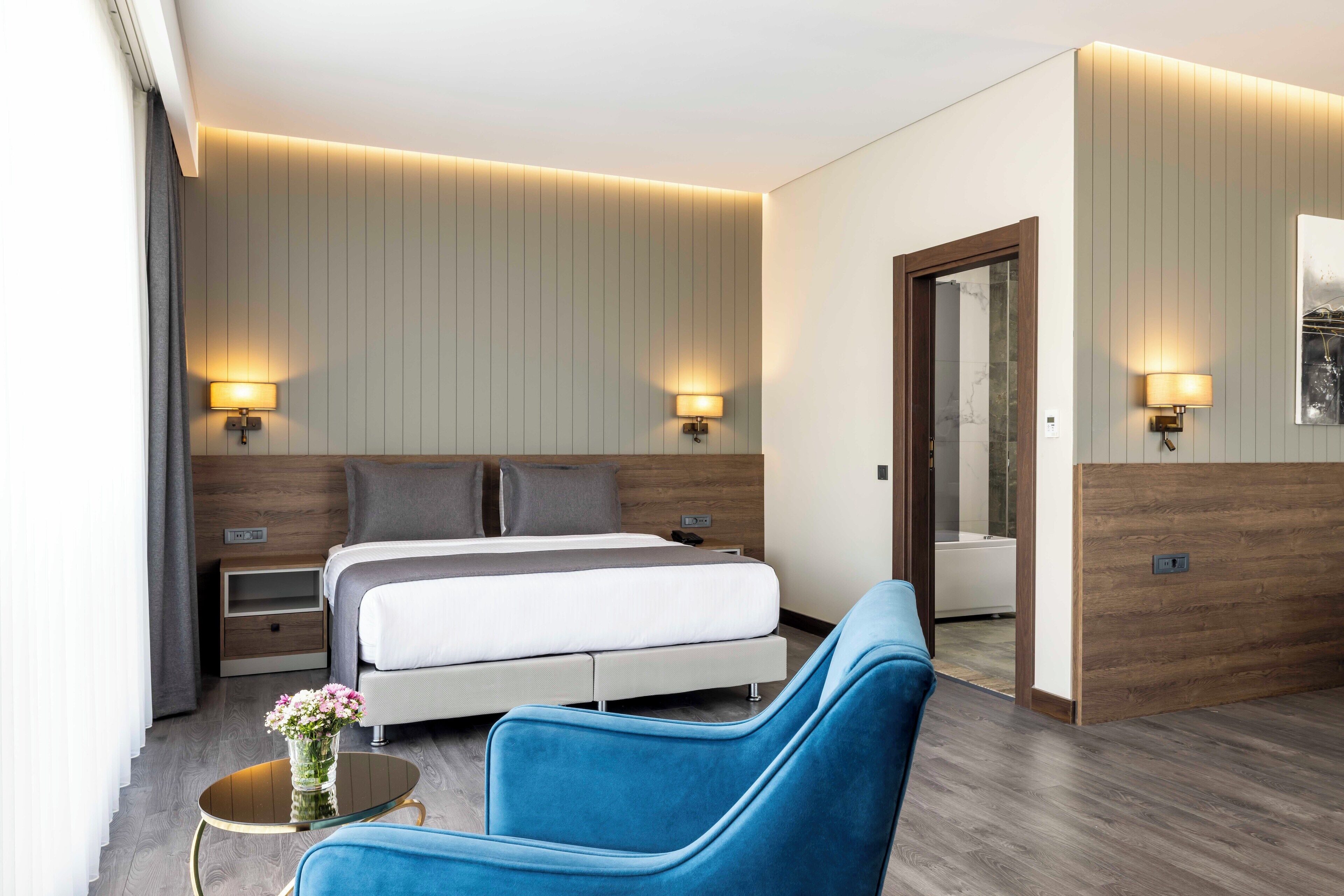 A modern hotel room with a bed, two bedside lamps, a small table with a vase, and a blue armchair.