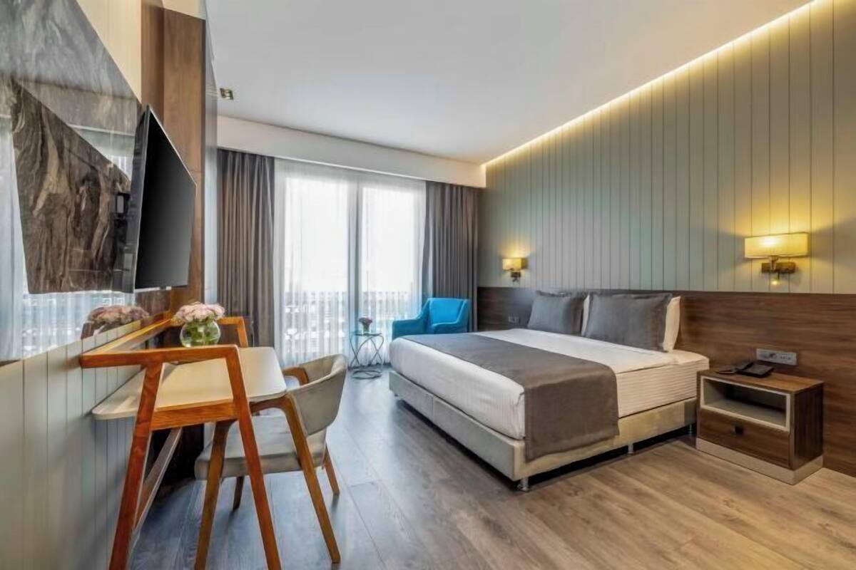 A modern hotel room with a bed, bedside table, desk, chair, and a TV mounted on the wall.