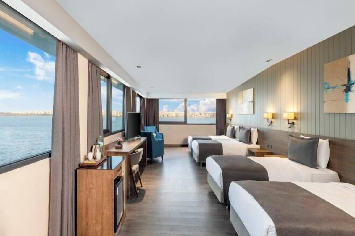 A hotel room with a large window, a desk with a chair, two beds, and a television.