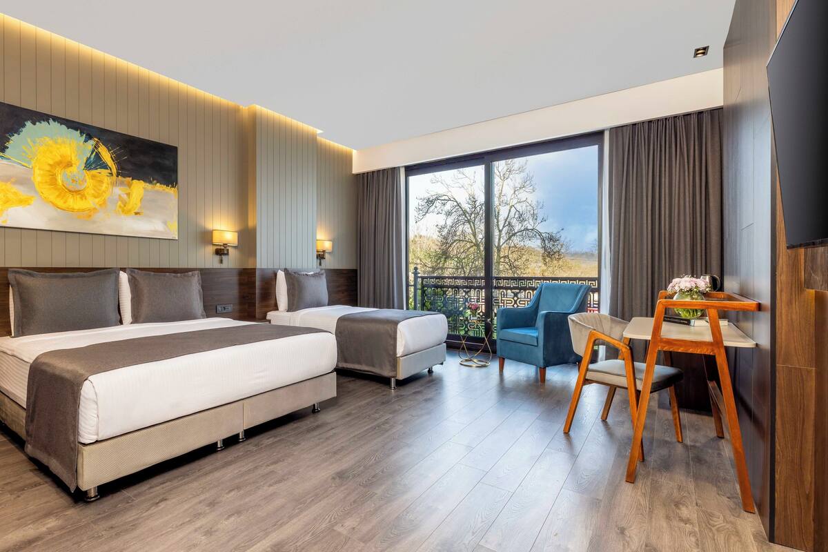 A hotel room with a large bed, a smaller bed, a desk with a chair, and a balcony with a view.