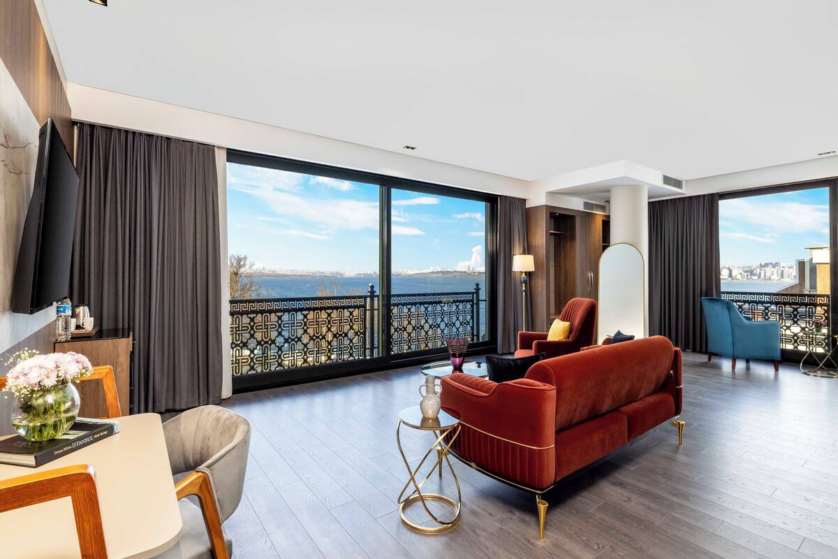 A modern hotel room with a large balcony, a red sofa, a dining area, and a view of the city.