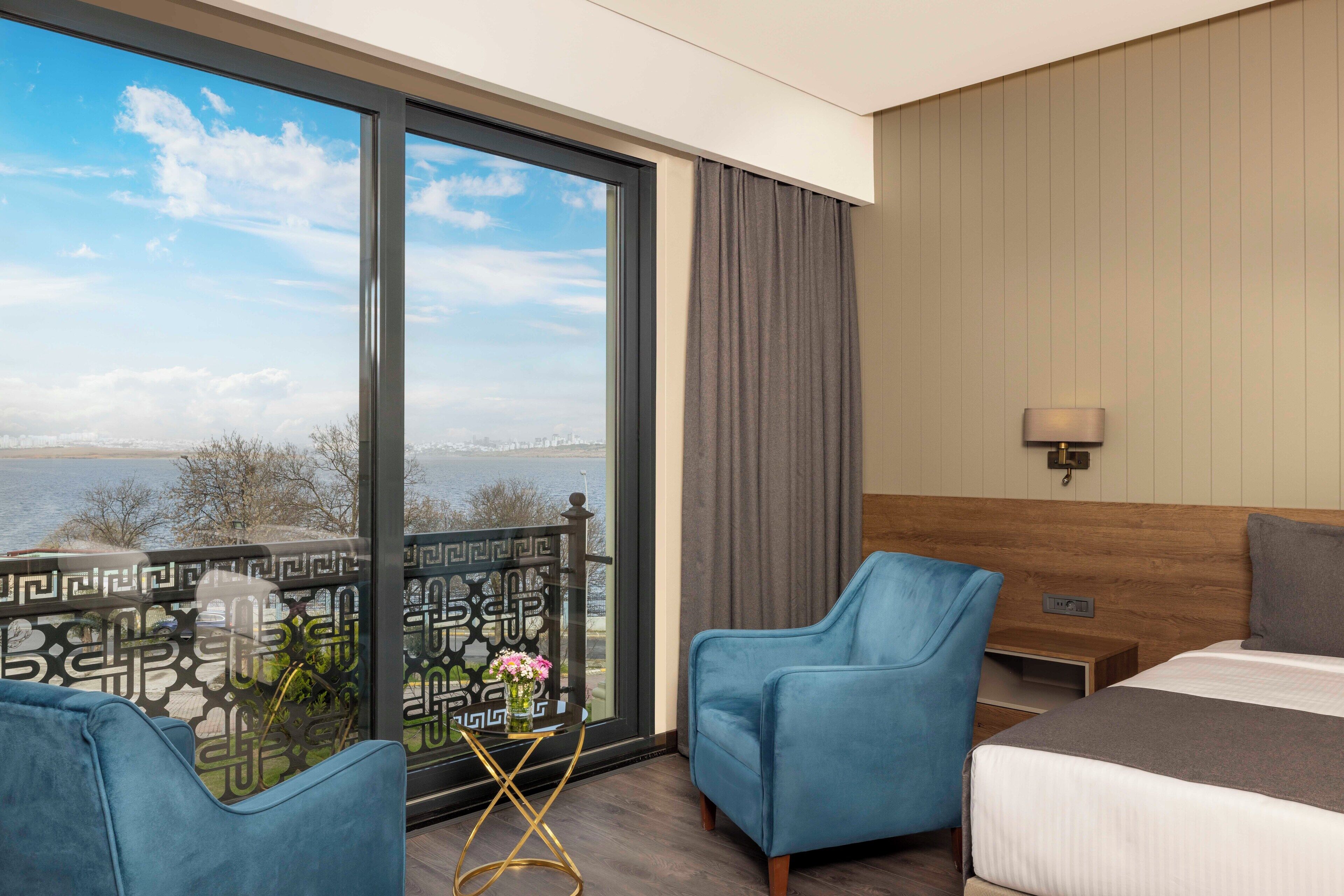 A hotel room with a large window, a bed, a blue armchair, a small table with a vase, and a nightstand.