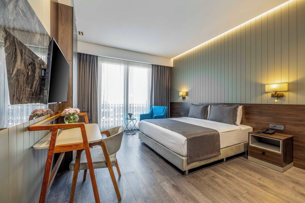 A modern hotel room with a large bed, a desk with a chair, a TV, and a nightstand with a phone.