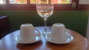 A table with two white cups and saucers, a clear glass vase with a plant, and a window with a view of greenery and a red building outside. - Serenity By Nature (Carrillos)