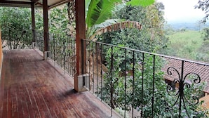 A covered balcony with a wooden floor and metal railing, overlooking a lush garden and a hilly landscape. - Serenity By Nature (Carrillos)